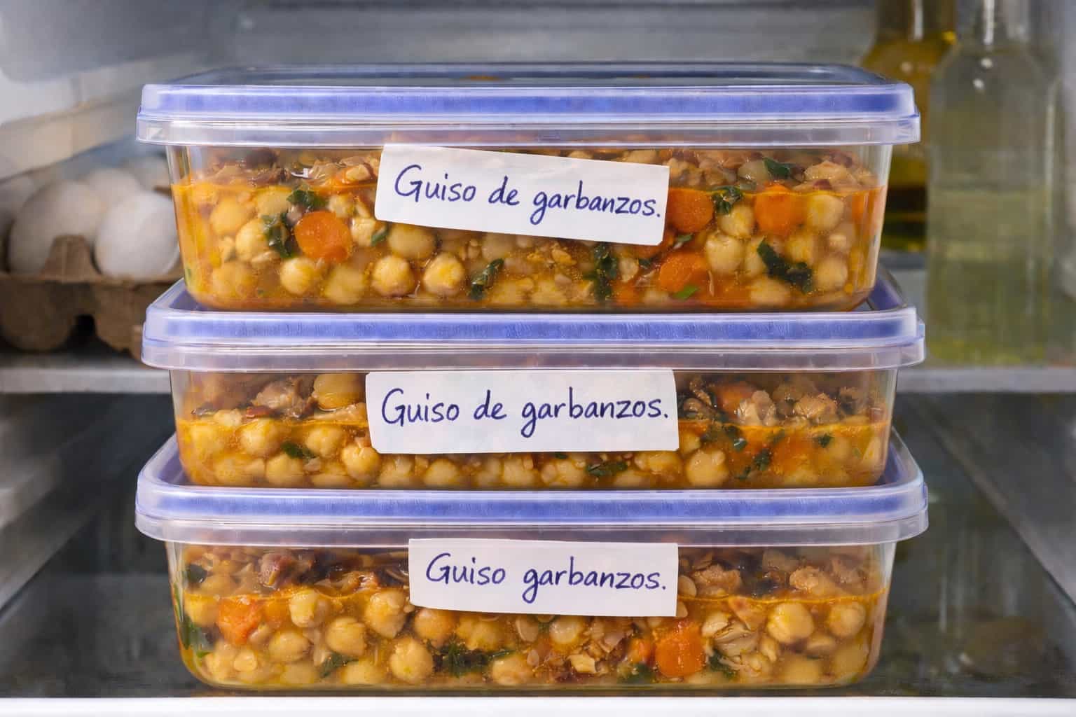 Tres t&aacute;peres con guiso de garbanzos apilados en una balda de nevera, con etiquetas de papel escritas a mano y ligeramente torcidas.