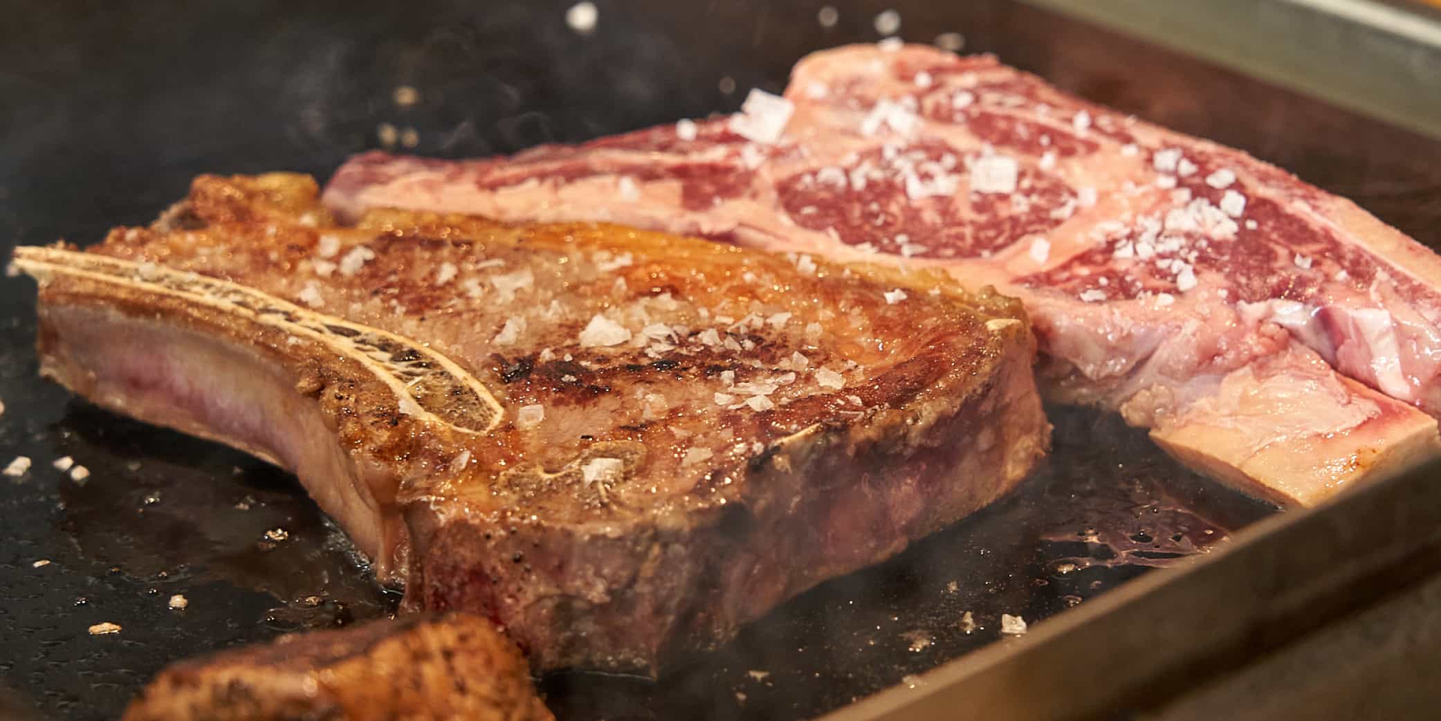 Primer plano de carne de vacuno cocin&aacute;ndose en plancha muy caliente, con sal en escamas y vapor visible. Imagen ideal para contenidos sobre cortes premium, sellado y t&eacute;cnicas de cocci&oacute;n.